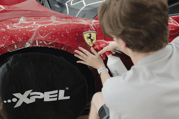 A technician applies a clear paint protection film to the front fender of a red Ferrari, using a spray bottle to smooth out the film near the iconic Ferrari emblem. The car's wheel is covered with a black XPEL-branded cover.