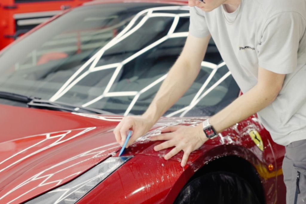 A technician smooths a freshly applied paint protection film over the hood and headlight of a red Ferrari using a blue squeegee tool. The vehicle's glossy finish reflects bright ceiling lights, emphasizing the precision of the application.