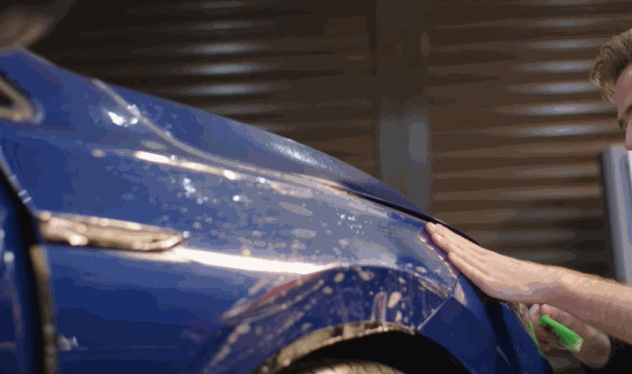 Technician smoothing clear paint protection film over a blue car fender to shield the paint from chips and scratches.