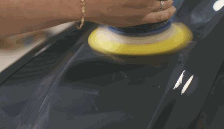 A machine polisher with a yellow pad works across a dark car panel to refine the surface before ceramic coating is applied. The paint reflects light smoothly as imperfections are removed during preparation.