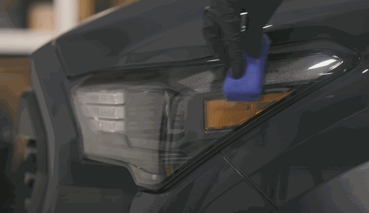 Close up of a dark vehicle headlight as a gloved hand uses a blue applicator pad to apply ceramic coating along the edge of the panel. The coating enhances gloss while adding a protective layer to the surface.