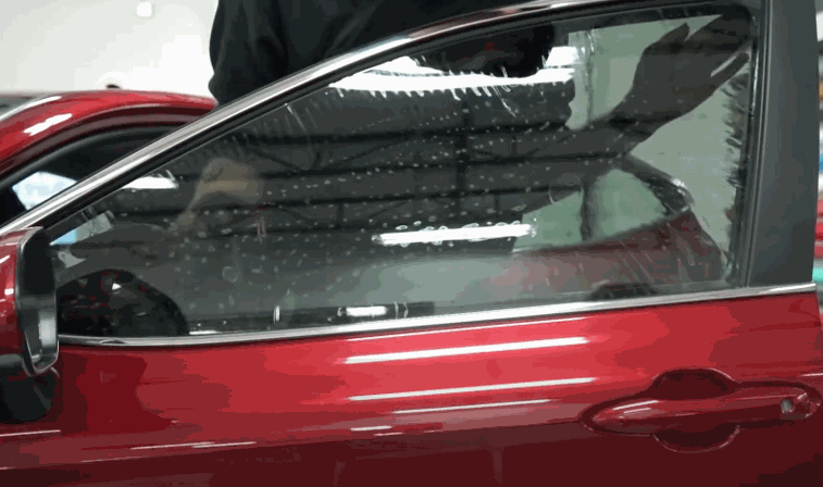 A red car door with window tint being installed from the inside as water droplets cover the glass. Overhead lights reflect across the surface while the film is being positioned evenly.